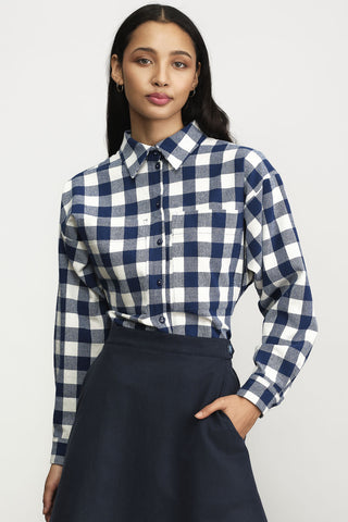 A woman modeling a long-sleeve button-up shirt with a navy and white check pattern, paired with a dark navy skirt, against a light gray studio background.