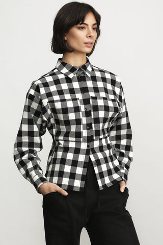 A woman wearing a black and white buffalo check plaid long-sleeve button-up shirt with black pants, standing against a light gray studio background.