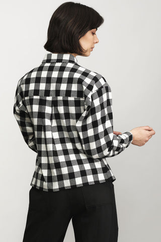 A woman shown from the back wearing a black and white checkered long-sleeve button-up shirt and black trousers, photographed against a plain light gray studio background.
