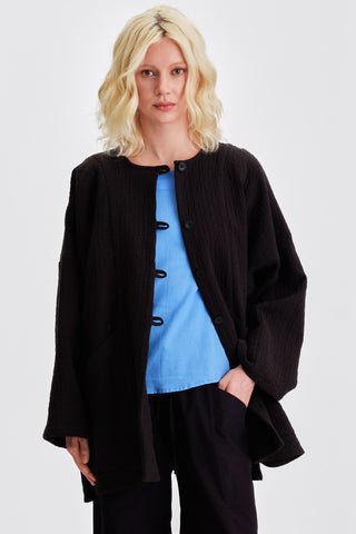 Woman in front of white background wearing black kimono style jacket over blue blouse and black pants. 