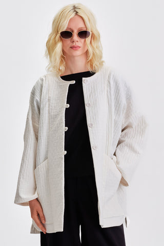 Woman in front of white background wearing ivory kimono style jacket over black blouse and pants. 