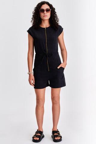 Woman in front of white background wearing sunglasses, black short zip up jumpsuit and black sandals. 