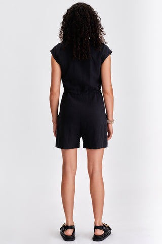 Back view of woman in front of white background wearing black short zip up jumpsuit and black sandals. 
