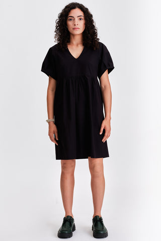 Woman in front of white background wearing black v-neck babydoll style dress with green leather lace-up shoes. 