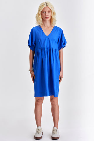 Woman in front of white background wearing vivid blue v-neck babydoll style dress with white sneakers. 