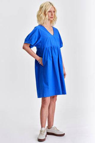 Woman in front of white background wearing vivid blue v-neck babydoll style dress with white sneakers. 