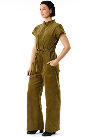 Side view of woman standing in front of white background wearing moss green corduroy Anath Jumpsuit by Jennifer Glasgow, featuring brass zipper, short sleeves and self-tie belt. 