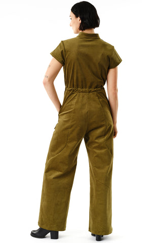 Back view of woman standing in front of white background wearing moss green corduroy Anath Jumpsuit by Jennifer Glasgow, featuring brass zipper, short sleeves and self-tie belt. 