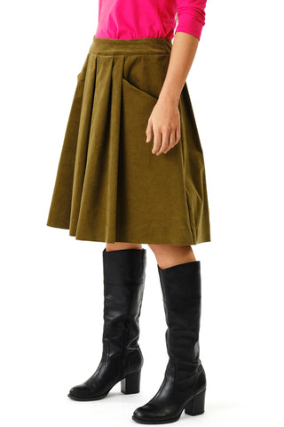 Close-up of woman against white background wearing olive green corduroy knee length Musica skirt by Jennifer Glasgow and black boots. 