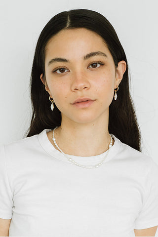 A woman in front of white background wearing white t-shirt, silver hoop earrings with freshwater pearl dangling charms and pearl necklace. 