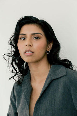 Woman in front of white background wearing grey collared shirt and small wavy sterling silver hoop earrings. 