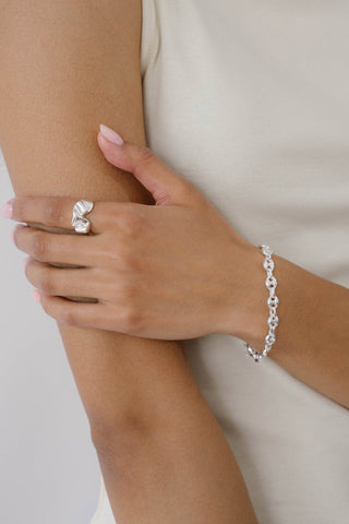 Close-up of a hand wearing a wavy silver ring and puffy mariner chain bracelet on a neutral background