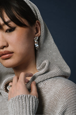 Close-up of a model wearing a chunky knit light gray hooded sweater with wavy silver earrings. 
