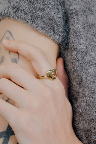 Gold heart-shaped ring with a red gemstone on a person's finger against a textured gray background.