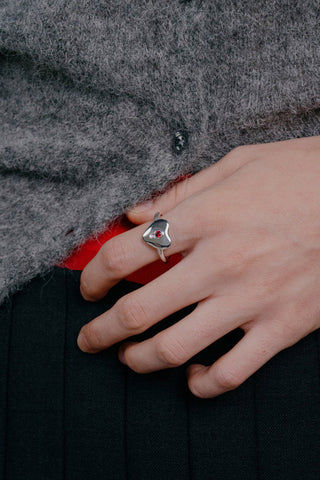 Hand wearing a silver heart shape ring with a ruby gem on a gray fabric background