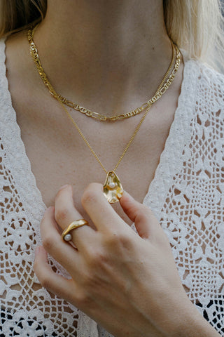 A close-up of a person wearing layered gold necklaces, including a textured chain necklace and an oyster shaped pendant necklace. 