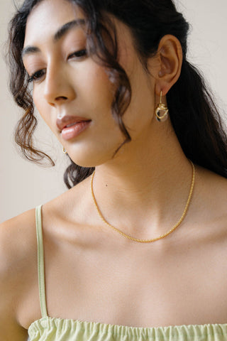 A close-up of a woman wearing a delicate gold chain necklace and matching gold hoop earrings, paired with a light green spaghetti-strap top. 