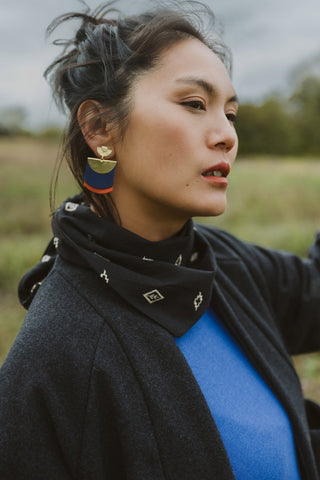 A woman outdoors wearing a blackscarf with small white geometric & floral patterns, a black cardigan over a bright blue top, and a gold and blue geometric earring.