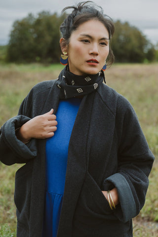 A woman standing outdoors in a grassy field wearing a dark charcoal coat over a bright blue dress, accessorized with a black patterned scarf and gold jewelry.
