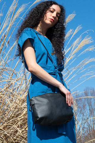 A woman with long curly dark hair wearing a blue dress and carrying a black leather shoulder bag, standing outdoors against a blue sky and dry reeds.