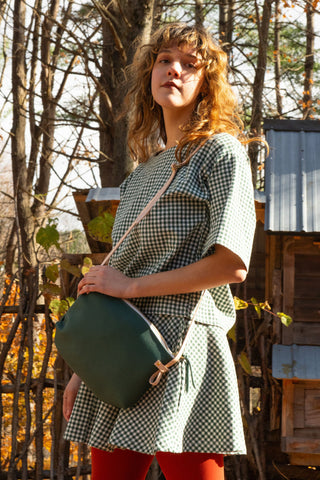 A woman wearing a green and white gingham dress and red tights, carrying a dark green crossbody bag with a beige strap, standing outdoors in front of trees and wooden structures.