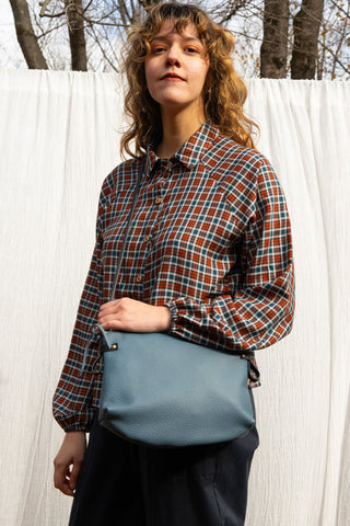 A woman with curly light brown hair stands outdoors in front of a white curtain, wearing a rust red, green, and blue plaid button-up shirt and dark trousers, while holding a dusty blue textured leather shoulder bag.