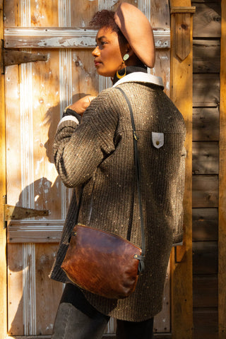 A woman wearing a brown textured tweed jacket with a white collar, a tan hat, and a brown leather shoulder bag, standing in front of a rustic wooden door.