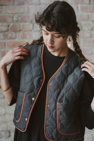 A woman wearing a black denim quilted vest with brown trim over a black top, posing in front of a brick wall.