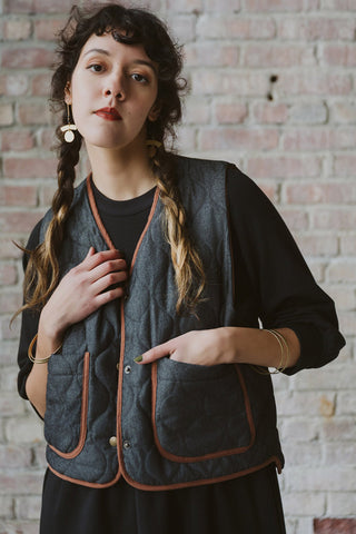 A woman wearing a black denim quilted vest with brown trim over a black top, posing in front of a brick wall.