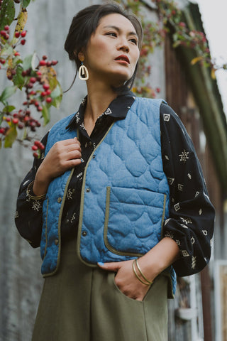 A woman wearing a blue denim quilted vest with olive trim over a black blouse and olive pants, posing in front of a wood panelled wall. 