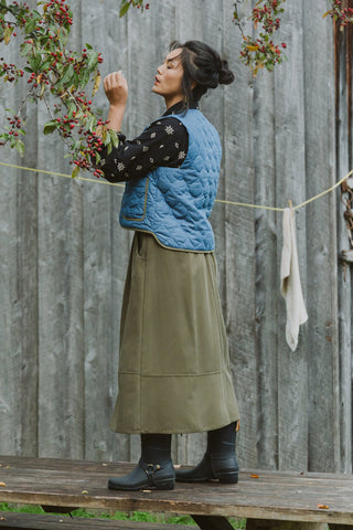 A woman stands on a wooden bench outdoors in front of a weathered gray barn wall, wearing a blue denim quilted vest over a black long-sleeve bloue, long olive green skirt, and black ankle boots.