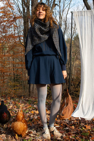 Women in forest in autumn wearing blue check scarf over navy dress holding brown leather bag. 