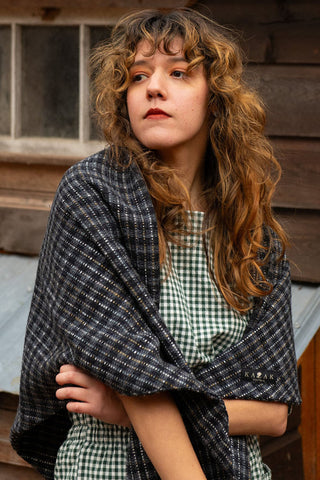 Women in front of old wood building wearing grey check scarf over gingham jumpsuit. 