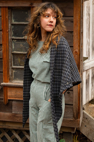 Women in front of old wood building wearing grey check scarf over gingham jumpsuit. 