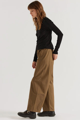 A woman modeling a black long-sleeve top and wide-leg brown pants, standing in profile against a plain light gray studio background.
