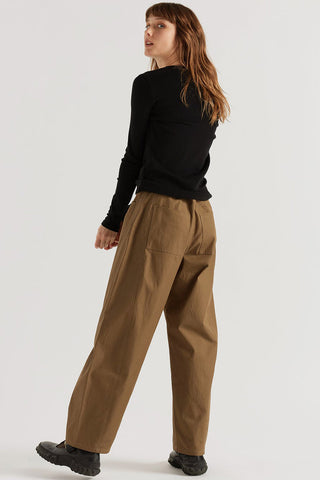 A woman wearing wide-leg brown pants, a black long-sleeve top, and black shoes, photographed from the back against a plain light gray studio background.
