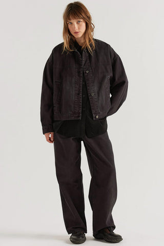 A model wearing a dark washed denim jacket, black shirt, and wide-leg dark trousers, standing against a light gray studio background.