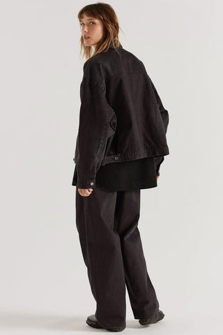 A model shown from the back wearing a black oversized denim jacket, a black shirt, and black wide-leg trousers against a light gray studio background.