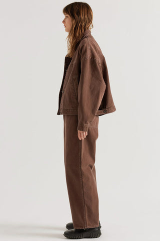 A model shown from the side profile wearing a brown oversized denim jacket and matching brown wide-leg pants against a plain light gray studio background.