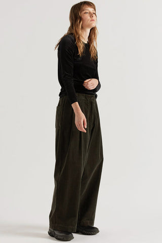 A woman modeling a black long-sleeve top and dark olive green / brown ballon leg pants against a plain light gray studio background.