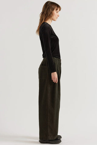 A woman stands in profile wearing a black long-sleeve top and dark olive green / brown wide-leg pants against a plain light gray studio background.