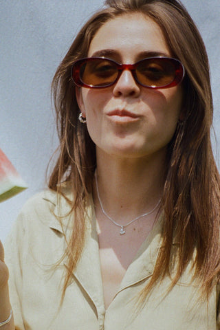 Woman wearing sunglasses, green button up shirt and silver heart shaped pendant on chain. 