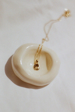 Gold pebble pendant necklace in cream ceramic bowl on white background. 