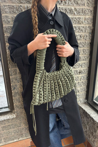 Woman standing in front of a brick wall wearing a black trench coat and holding an olive green chunky crocheted shoulder bag. 