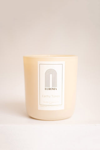 Candle in beige glass container with white label that reads "Luroma - Earthy Tones". Photographed in front of beige background. 