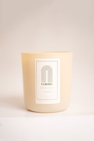 Candle in beige glass container with white label that reads "Luroma - White Tea + Thyme". Photographed in front of beige background. 