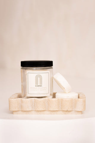 A glass jar and two shower steamers on beige soap dish, in front of beige background. 