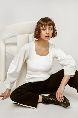 Woman sitting on floor in front of white chair, wearing black pants and white square neck Blair top by Marigold. 