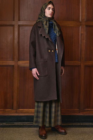 A model stands against a dark wood-paneled wall wearing a long brown wool coat with scalloped edges, a green and brown plaid headscarf, a blue patterned sweater, a matching plaid skirt, and brown leather boots.