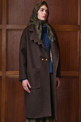 A model wearing a dark brown wool coat with scalloped edges, a green plaid headscarf, and a blue patterned top, standing in front of a dark wood paneled wall.